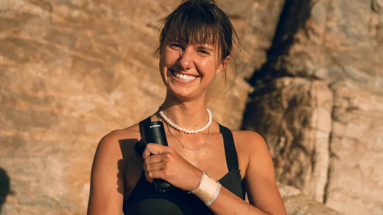 Femme souriante tenant un pistolet de massage type Theragun ou Hypervolt dans un cadre ensoleillé